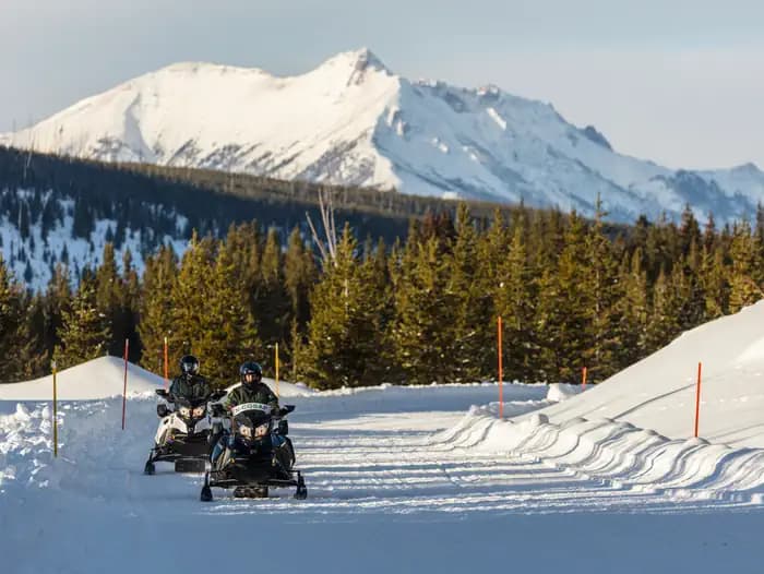 Two people on snowmobiles travel on a groomed road in Yellowstone National Park with Electric Peak in the background.