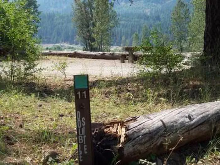 Site marker eleven in foreground; picnic table in a flat gravel area with bushes encircled with log barrier, river valley and conifer covered hills in background.