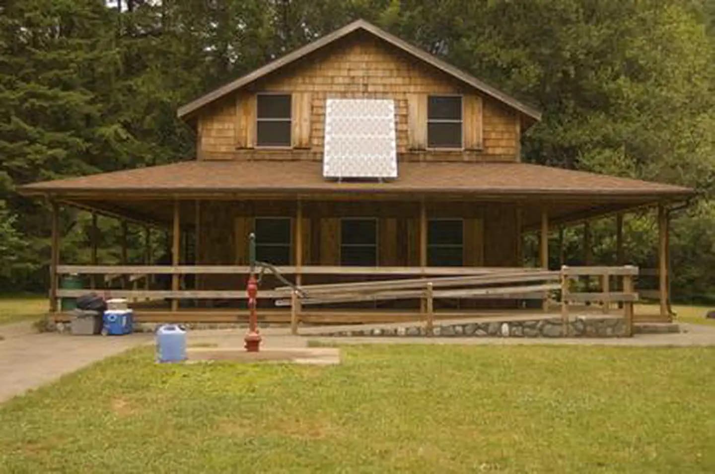 House with shingle siding,  solar panel on the roof of wrap around covered porch, hand pump well on the lawn, and ramp leading up to porch