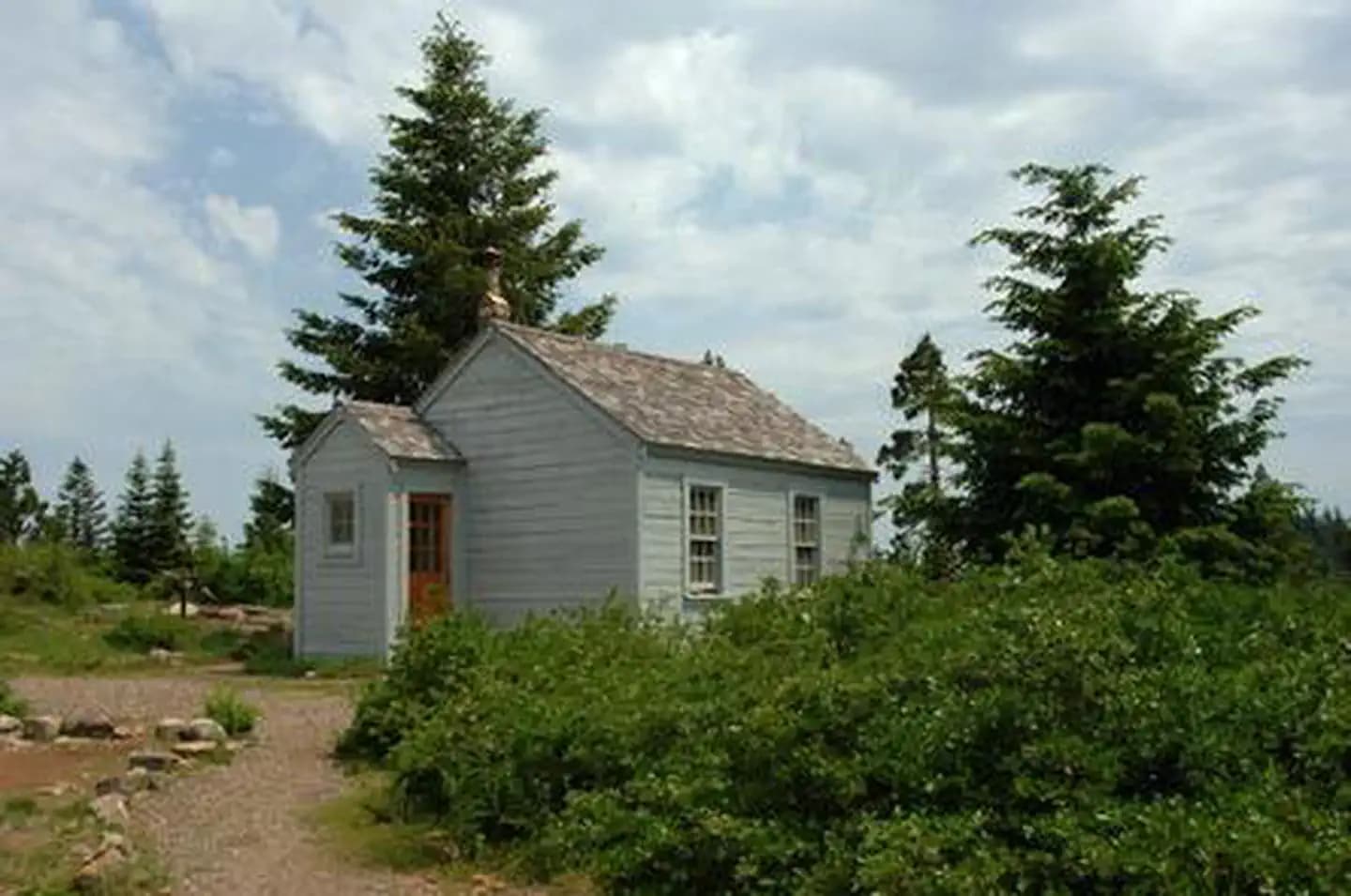 Green shrubs next to a gravel path leading to a small gray cabin with a partly cloudy sky and two tall conifer trees as a backdrop.   