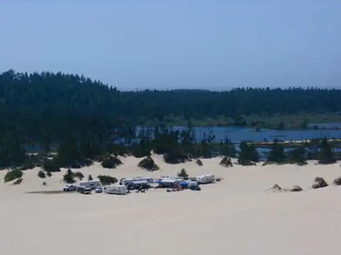 Group of trailers and trucks on a large expanse of sand near a conifer encircled lake under a blue sky.