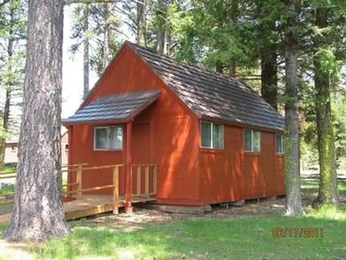 Small, orange-red cabin with ramp to front door in sun dappled conifer forest.