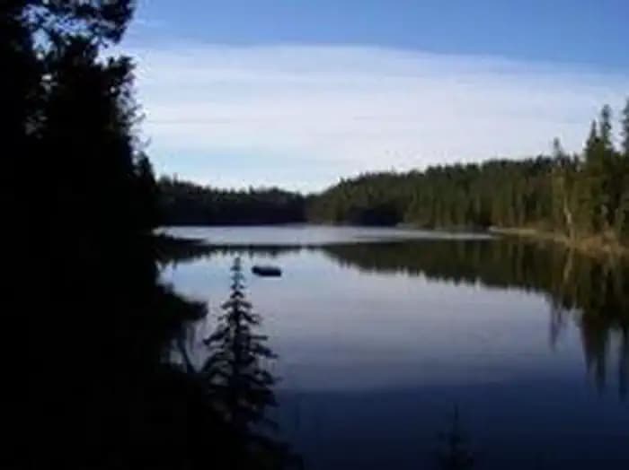 Glassy lake reflecting conifer lined shore, blue sky and cloud.