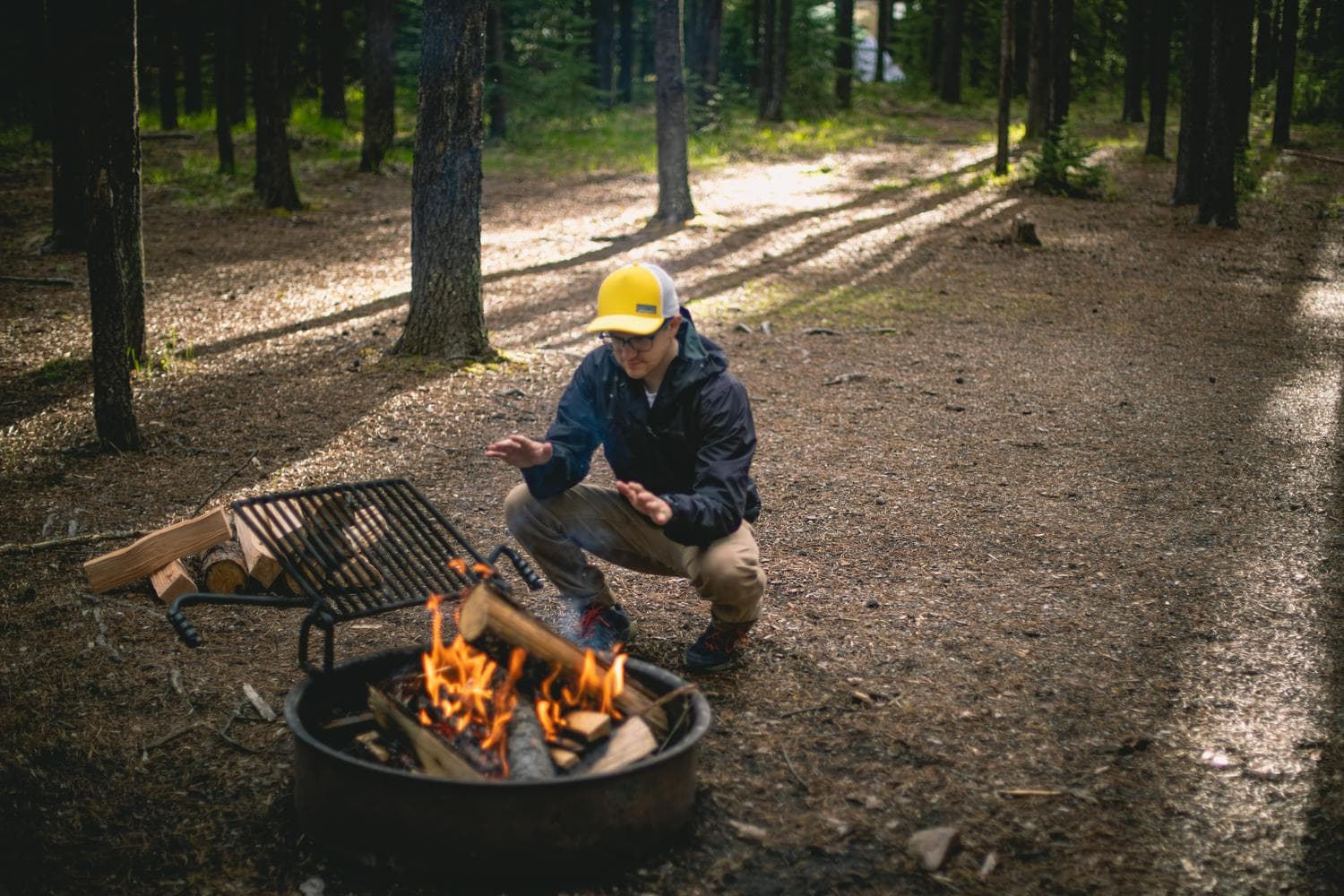 A camper wearing a bright hat warms their hands while squatting near a campfire in a forest clearing.