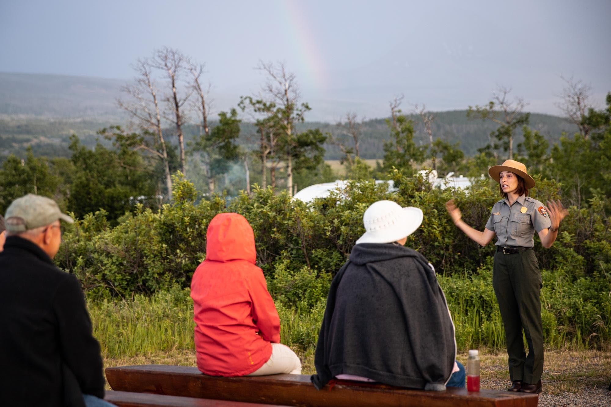 A park ranger in uniform stands in front of a crowd and gestures with their hands while giving an evening program to a group of park visitors. A rainbow is faintly visible in the background.
