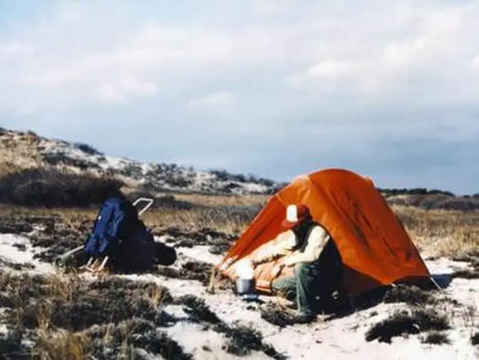 Backpacker cooking food in front of an orange tent with sand dunes in the background